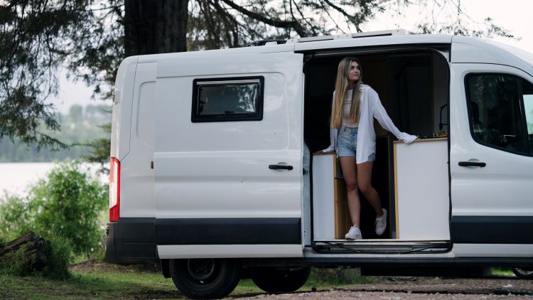 How to Prepare for Full-Time RV Living Woman full time rv living standing in doorway of a white camper van, looking out at the calm lake and surrounding forest
