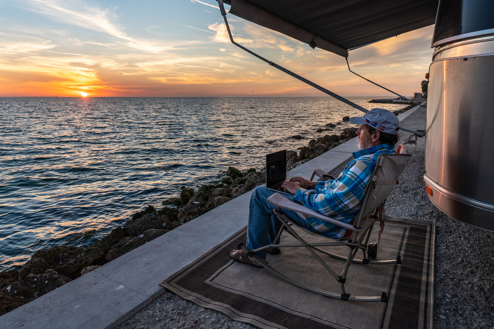 A man sitting on a chair by the ocean watching the sunset while working on his laptop computer remotely.