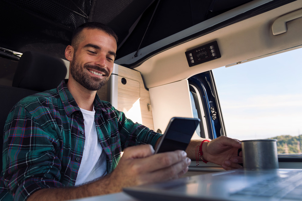smiling young man using his mobile phone and laptop from his camper van in the middle of nature.