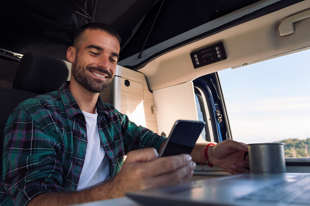 smiling young man using his RV wifi smiling young man using his mobile phone and laptop from his camper van in the middle of nature.