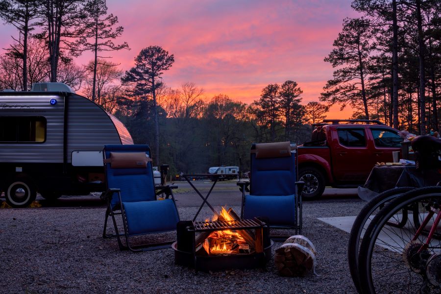 Campground at Sunset Two camp chairs sitting next to a fire. A travel trailer is visible in the background, with a pink sunset sky.