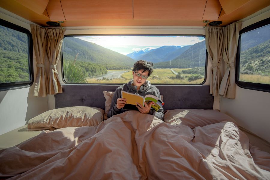 Young man in an rv bed reading a book with mountain scenery in the background
